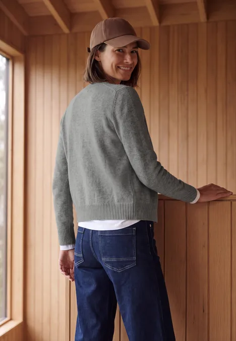 Eine Frau in grauem Pullover und Jeans, mit brauner Kappe, blickt lächelnd über die Schulter in einen Holzraum.