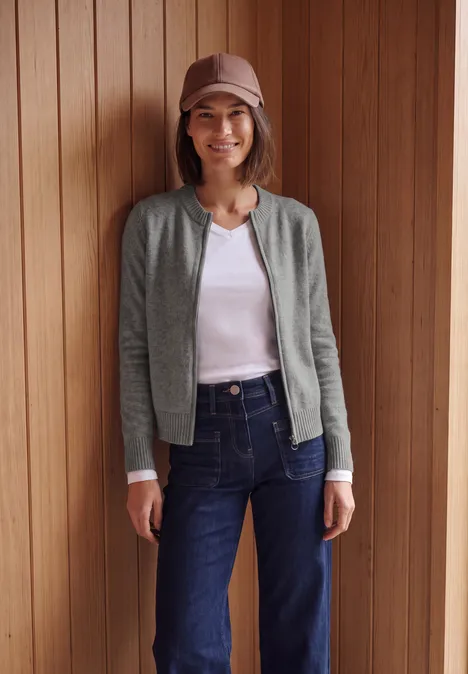 Frau vor Holzpaneelen, trägt Jeans, weißes Shirt, graue Jacke, Mütze, lächelt.