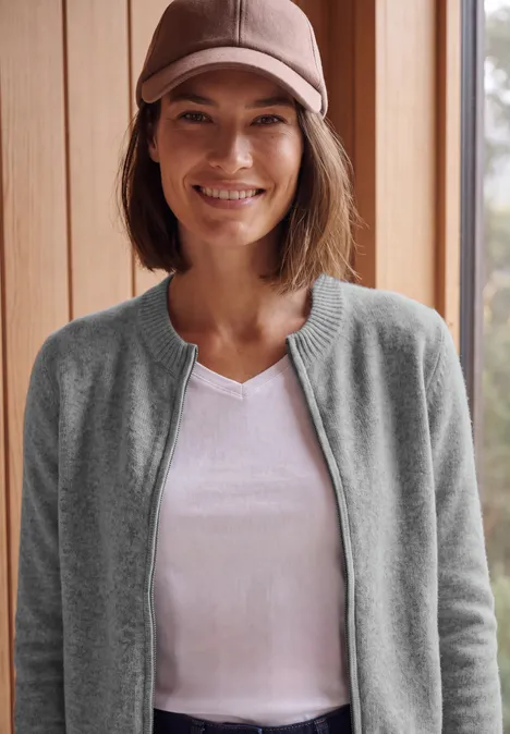 Eine lächelnde Frau mit Basecap, weißem T-Shirt und grauem Cardigan vor Holzwand.