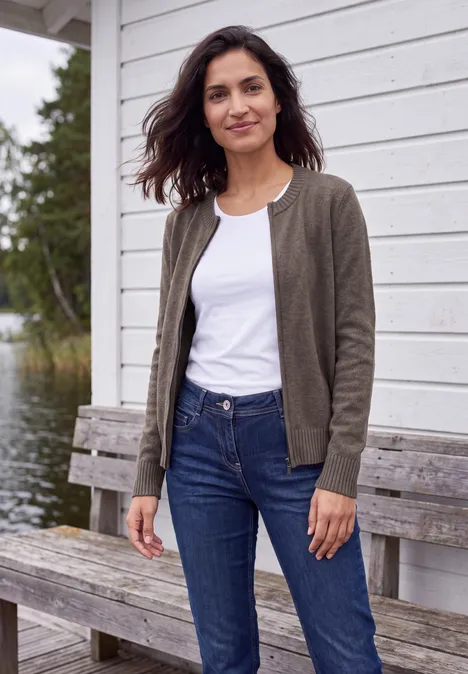 Langarm Cardigan mit Rundhals und Zipper mushroom taupe melange Eine Frau in Jeans, weißem T-Shirt und grauer Jacke steht lächelnd vor einer Holzhütte am See.