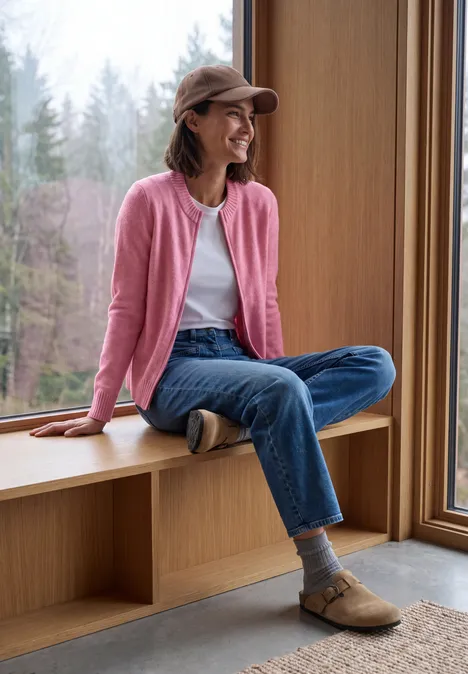Frau in rosa Jacke und Jeans sitzt lächelnd am Fenster und blickt in den Wald.