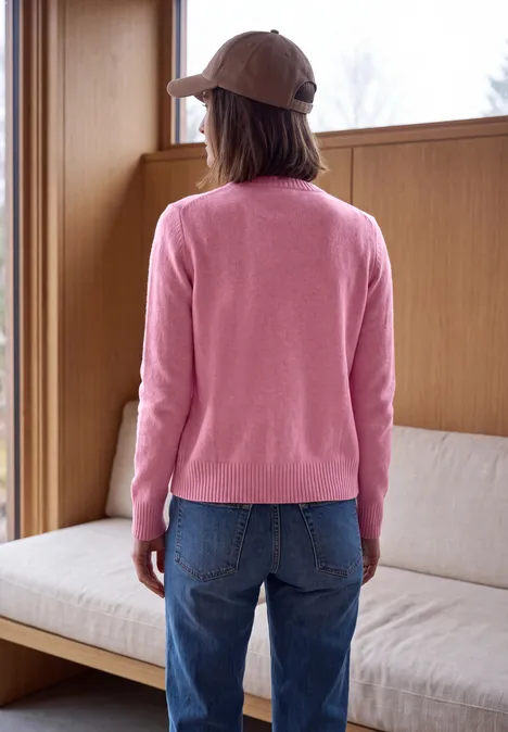 Eine Person von hinten mit rosa Pullover, Jeans und Baseballcap steht vor einem Sofa.
