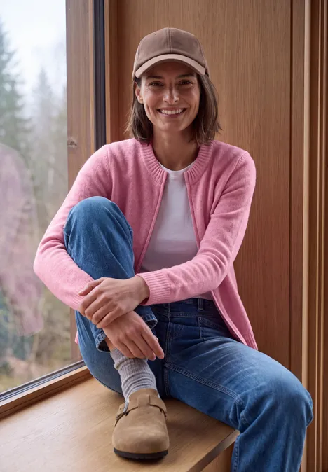 Eine lächelnde Frau in rosa Cardigan und Jeans sitzt auf einer Fensterbank.