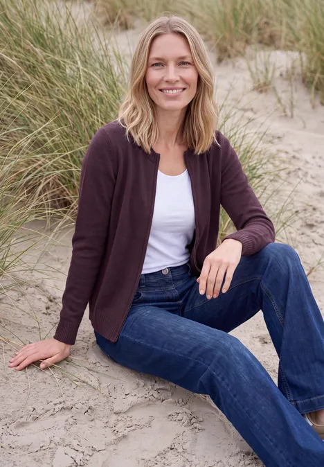 Langarm Cardigan mit Rundhals und Zipper eggplant red melange Frau sitzt entspannt im Stranddünensand, trägt Jeans, weißes Shirt und Strickjacke, lächelt.