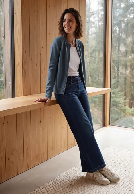 Langarm Cardigan mit Rundhals und Zipper cotton blue melange Frau in Jeans und Cardigan lehnt entspannt an einer Holzbank in modernem Raum mit großen Fenstern.