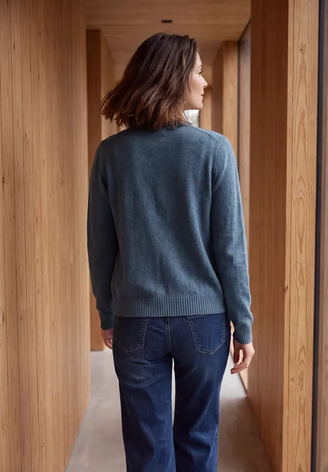 Langarm Cardigan mit Rundhals und Zipper cotton blue melange Frau im grauen Pullover und Jeans steht in einem hellen Holzflur, Rücken zur Kamera, Blick aus dem Fenster.