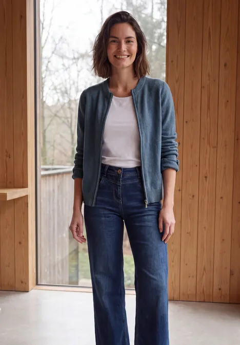 Langarm Cardigan mit Rundhals und Zipper cotton blue melange Junge Frau in Jeans und weißem Shirt, lächelnd und locker, vor Holzpaneelen und Fenster stehend.