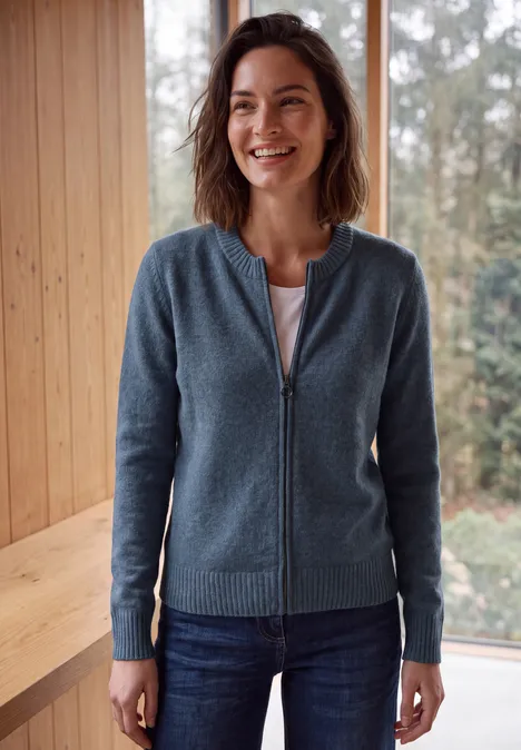 Langarm Cardigan mit Rundhals und Zipper cotton blue melange Eine lächelnde Frau in blauem Reißverschluss-Pullover und Jeans steht drinnen vor Holzverkleidung und Fenstern.
