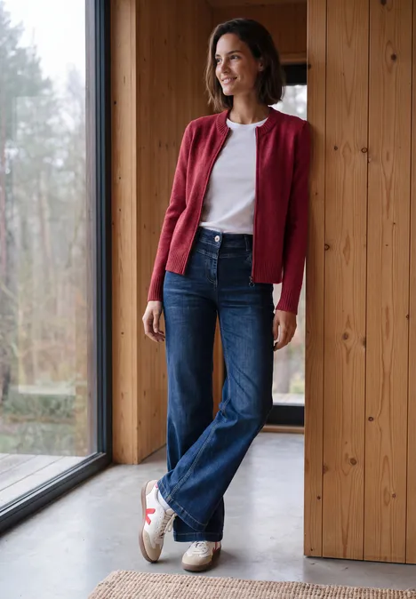 Langarm Cardigan mit Rundhals und Zipper rhubarb melange Frau in roter Strickjacke, weißem Shirt und Jeans lehnt entspannt am Fenster in modernem Holzhaus.