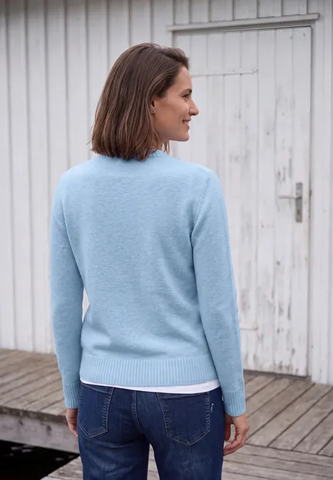 Frau von hinten in hellblauem Pullover und Jeans vor einer weißen Holztür auf Holzterrasse.