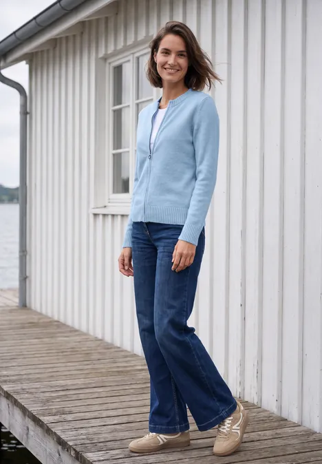 Frau lächelt auf Holzsteg am Wasser neben weißem Haus, trägt hellblaue Jacke, Jeans und bequeme Schuhe.