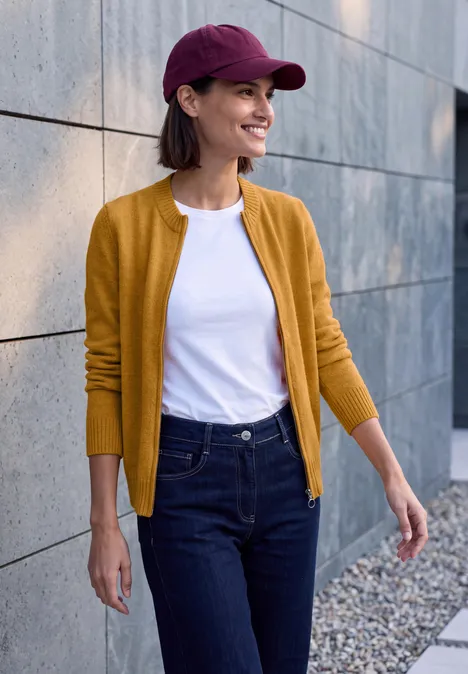 Frau in gelbem Cardigan, weißem T-Shirt und Jeans mit roter Kappe spaziert lächelnd an einer Steinmauer entlang.