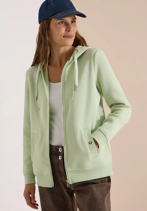 Frau in pastellgrüner Kapuzenjacke, weißem Top, brauner Hose und blauer Mütze lächelt seitlich.