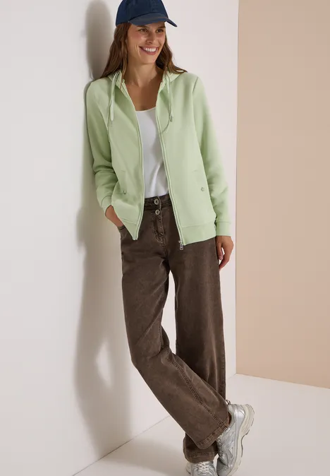 Junge Frau lehnt an Wand, trägt grünen Cardigan, weißes Top, braune Cordhose, weiße Sneaker und blaue Mütze.