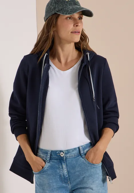 Frau in weißem T-Shirt, dunkelblauer Kapuzenjacke und Jeanscap, mit Händen in den Hosentaschen.