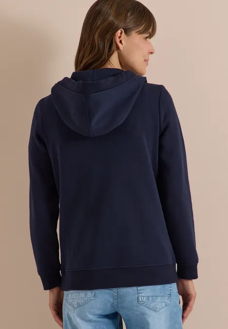 Frau von hinten in dunkelblauer Kapuzenjacke und hellen Jeans vor neutralem Hintergrund.