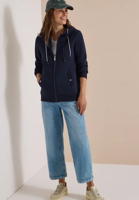 Frau trägt dunkelblaue Kapuzenjacke, graues Shirt, weite Jeans und Sneaker, lehnt lächelnd an der Wand.