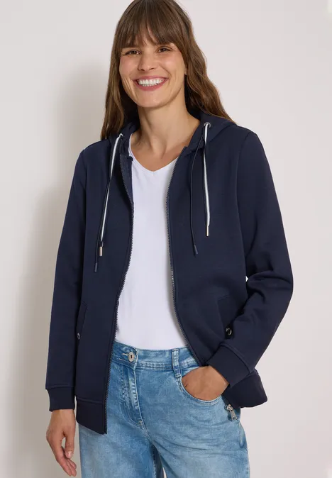 Junge Frau in dunkler Kapuzenjacke, weißem Shirt und Jeans, lächelnd, Hand in Tasche.