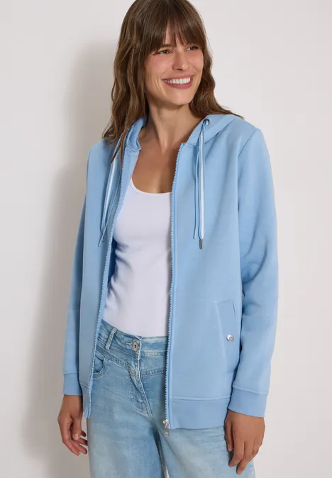 Eine lächelnde Frau trägt einen hellblauen Hoodie über einem weißen Top und Jeans.