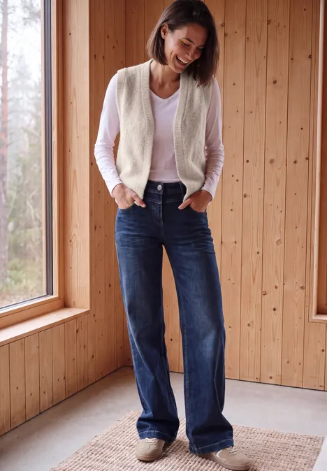 Offene Federgarn-Weste in Unifarbe cashmere beige Eine lächelnde Frau in Jeans und Weste steht entspannt vor einer Holzverkleidung am Fenster.
