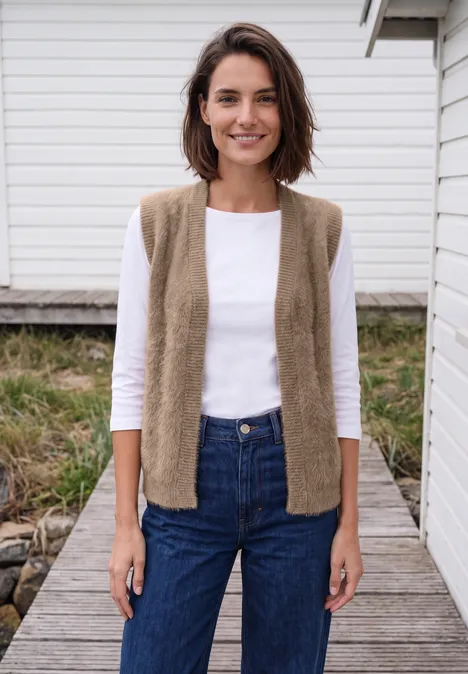 Eine Frau in Jeans, weißem Shirt und beigem Strickweste steht lächelnd auf einem Holzsteg.