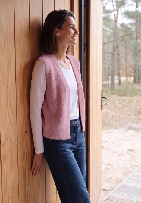 Open vest van veergaren in effen kleur velvet rose Eine Frau steht lächelnd in einer Holztüröffnung, trägt eine rosa Weste, weißen Pullover und Jeans. Wald im Hintergrund.