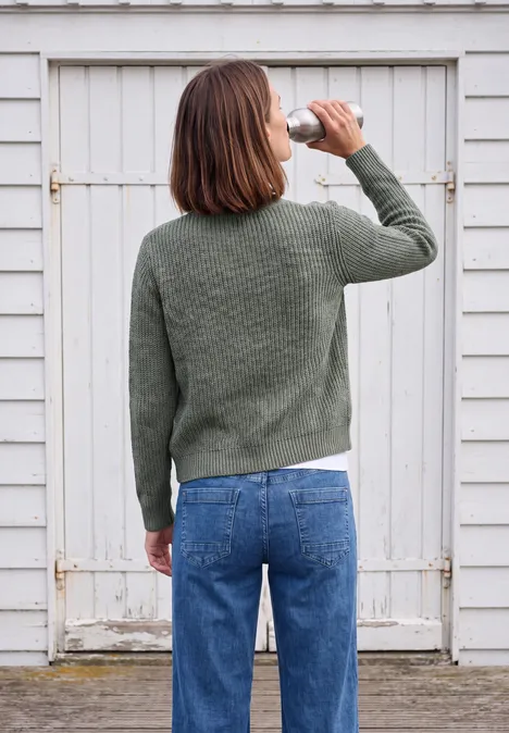 Une femme en pull vert et jean bleu boit dans une bouteille, vue de dos devant une porte en bois blanc.