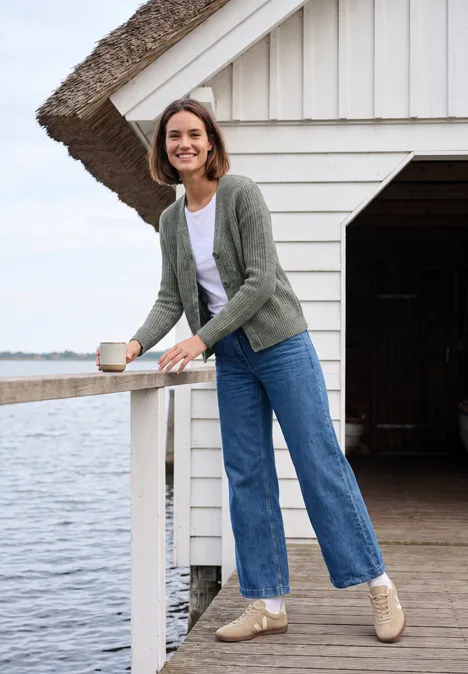 Femme appuyée sur une barrière d'un ponton au bord du lac, souriante, tasse à la main, jean et cardigan.