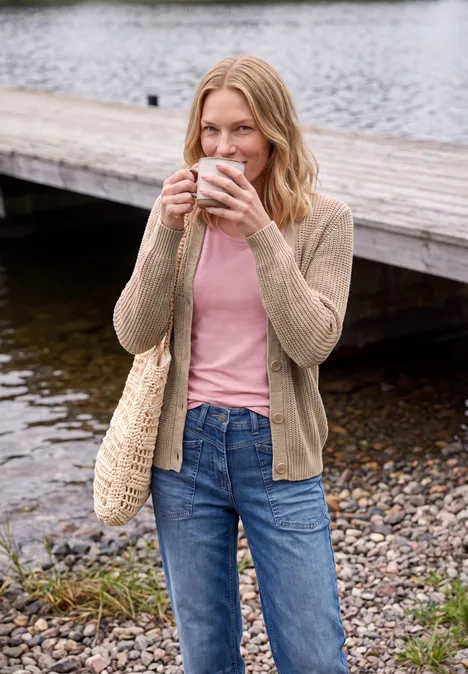 Femme assise sur une jetée en bois au bord d'un lac, tenant une tasse, en jean, t-shirt rose, gilet beige et sac en paille.