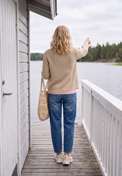 Femme debout sur une terrasse en bois, sac en osier, jean et pull, regardant un lac paisible.