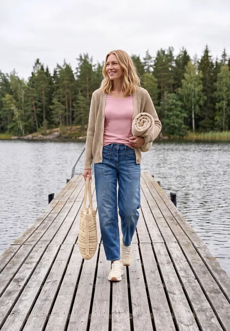 Une femme marche souriante sur une jetée en bois au bord du lac, tenant une couverture et un panier, entourée de forêt.