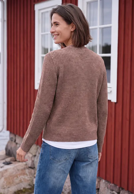 Chunky Cardigan mit V-Neck und Melange-Optik dark russet red melange Eine Frau mit braunem Pullover und Jeans steht lächelnd vor einem roten Holzhaus und schaut weg.