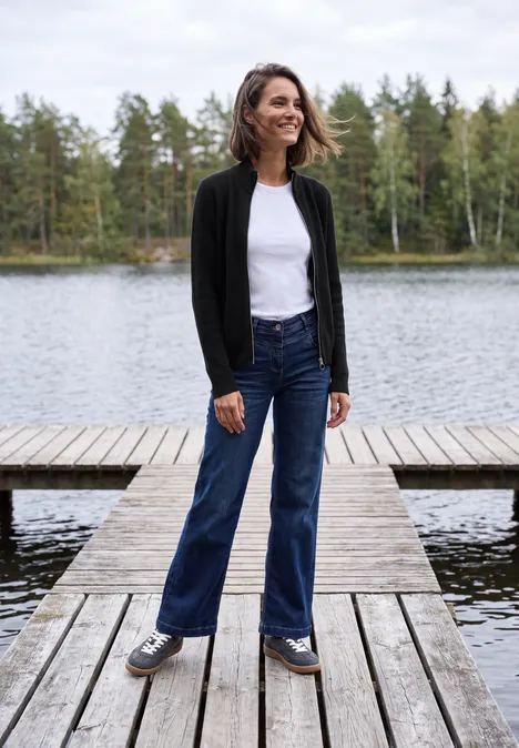 Cardigan mit Stehkragen und Zipper Black Eine Frau in Jeans und Jacke steht lächelnd auf einem Steg an einem See, umgeben von Wäldern.