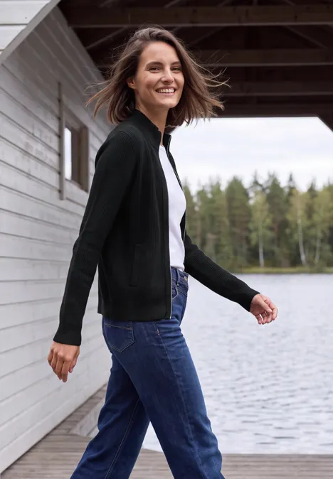 Cardigan mit Stehkragen und Zipper Black Frau in schwarzer Jacke und Jeans spaziert an einem Seeufer neben einer Holzhütte, lächelnd.