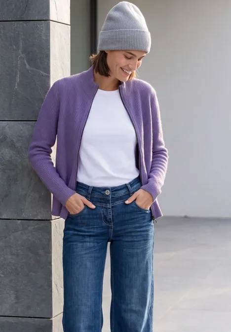 Cardigan mit Stehkragen und Zipper pastel lilac melange Frau in lila Strickjacke, weißem Shirt, Jeans und Mütze lehnt lächelnd an einer Mauer.