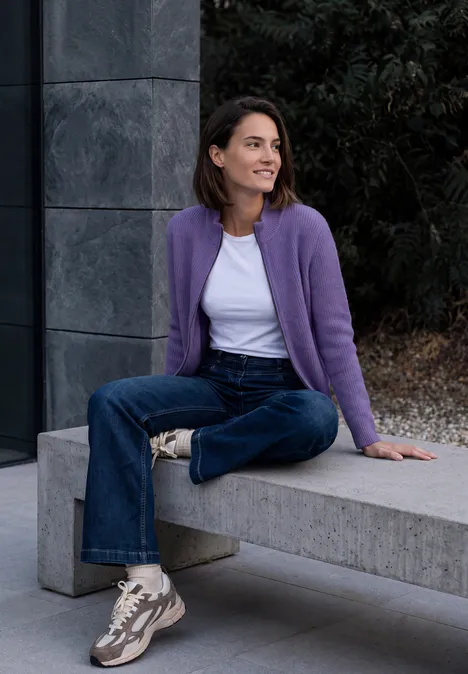 Cardigan mit Stehkragen und Zipper pastel lilac melange Eine Frau sitzt lächelnd auf einer Betonbank, trägt lila Strickjacke, weiße Bluse, Jeans und Sneaker.