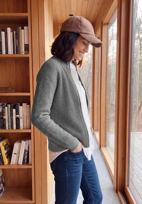 Cardigan zippé à col montant flint grey melange Frau in grauem Cardigan und Baseballcap steht entspannt am Fenster vor Bücherregal und blickt nach draußen.
