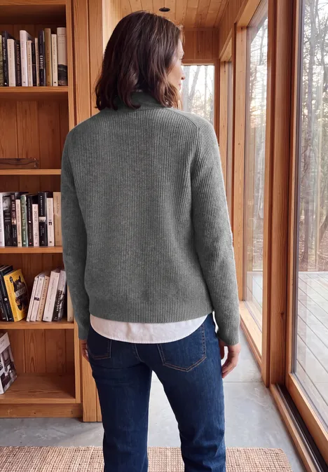 Cardigan zippé à col montant flint grey melange Person im grauen Pullover und Jeans steht vor Holzregal und großen Fenstern und blickt hinaus.
