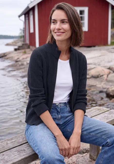 Cardigan mit Stehkragen und Zipper urban dark blue Eine Frau sitzt lächelnd auf einer Holzbank an einem Felsen am Wasser mit einem roten Haus im Hintergrund.