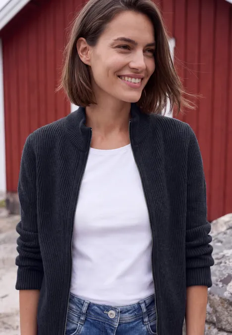 Cardigan mit Stehkragen und Zipper urban dark blue Eine lächelnde Frau trägt schwarzen Cardigan und weißes Shirt vor roter Holzwand.