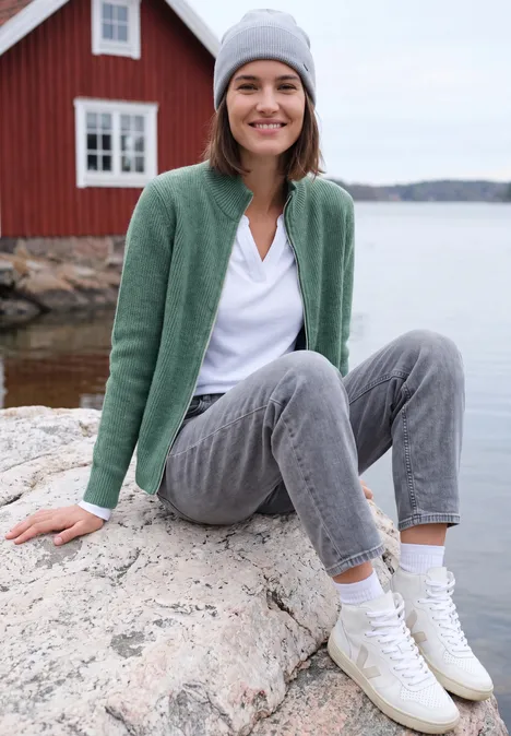 Cardigan mit Stehkragen und Zipper jewel green melange Junge Frau in grüner Strickjacke, grauer Hose und Mütze sitzt auf einem Felsen am Wasser vor rotem Haus.