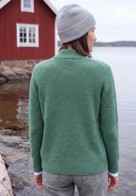 Cardigan mit Stehkragen und Zipper jewel green melange Eine Person steht im grünen Pullover und grauer Mütze mit Blick aufs Wasser vor einem roten Haus.