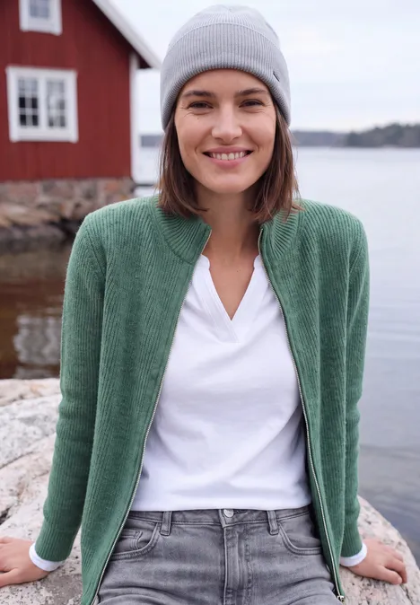 Cardigan mit Stehkragen und Zipper jewel green melange Frau mit grauer Mütze, grünem Cardigan und weißem Shirt steht entspannt am Ufer vor rotem Haus.
