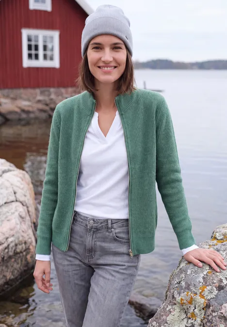 Cardigan mit Stehkragen und Zipper jewel green melange Eine Frau in grauer Mütze und grüner Strickjacke steht am felsigen Ufer vor einem roten Haus.