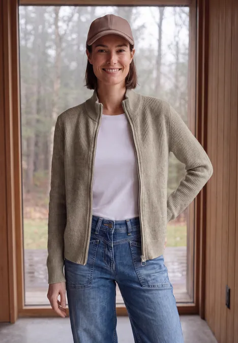 Femme souriante portant une casquette, une veste légère vert clair, un t-shirt blanc et un jean, debout dans une porte.