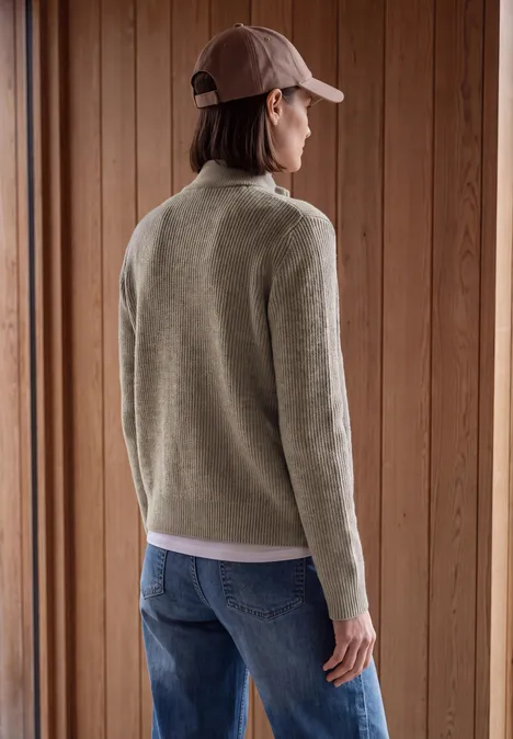Femme de dos en jean bleu, pull beige et casquette rose devant un mur en bois.