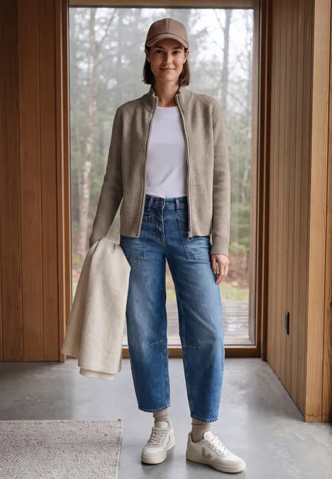 Femme décontractée en casquette, veste gris clair, t-shirt blanc, jean et baskets, tenant un manteau beige