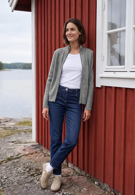 Cardigan mit Stehkragen und Zipper sporty grey melange Frau lehnt entspannt an einem rot gestrichenen Haus am See, trägt Jeans, weißes Shirt und graue Strickjacke.