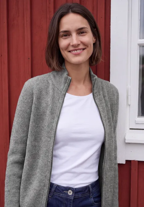Cardigan mit Stehkragen und Zipper sporty grey melange Eine lächelnde Frau in weißem Shirt und grauer Jacke steht vor einer roten Holzfassade mit Fenster.