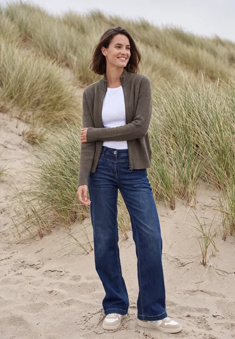 Cardigan mit Stehkragen und Zipper mushroom taupe melange Frau im weißen Shirt, grüner Strickjacke und blauen Jeans steht lächelnd in Dünengras am Strand.